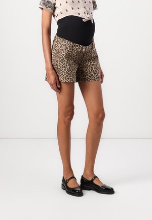 Femme portant un short de maternité imprimé léopard, une chemise légère à motifs nouée et des chaussures Mary Jane noires, debout sur un fond uni.
