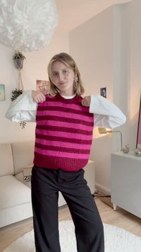 Pleteni brezrokavnik s horizontalnimi črtami v temno rdeči in roza barvi. Okr round-neck, teksturiran vzorec, nošen preko bele majice z dolgimi rokavi, v kombinaciji s črnimi hlačami.