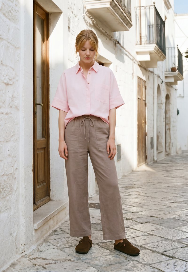 Femme en chemise rose à manches courtes et pantalon beige à cordon debout dans une rue pavée près de bâtiments en pierre blanche.