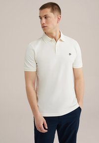 Krémové polo tričko s krátkými rukávy, jemnou texturou a vyšitým logem na hrudi. Nosené s tmavě modrými kalhotami.