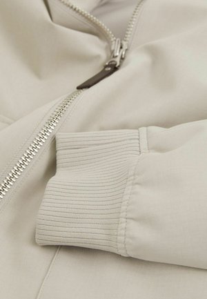 Giacca zip-up beige chiaro con una finitura liscia, caratterizzata da una cerniera argentata e polsini a coste. Il tessuto ha una consistenza morbida e una leggera lucentezza.