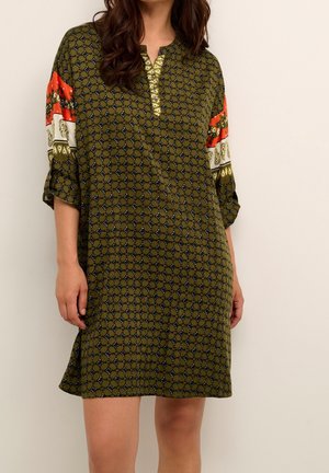 Vrouw draagt een olijfgroene, knie-lange jurk met patroon, met oranje, witte en gouden details op de mouwen en de halslijn.