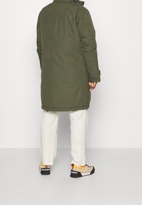 Lång olivgrön kappa med en slät yta, utrustad med hög krage, fickor på sidan och en logodetalj på baksidan. Bärs med ljusa byxor och sneakers.