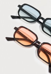 Δύο ζευγάρια ορθογώνιων γυαλιών ηλίου με μαύρα πλαίσια, το ένα με μπλε φακούς και το άλλο με πορτοκαλί φακούς, πάνω σε λευκή επιφάνεια.