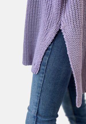 Lavendelfarbene Stricktunika mit strukturiertem Finish, mit seitlichem Schlitz, getragen über dunkelblauen, eng anliegenden Jeans.
