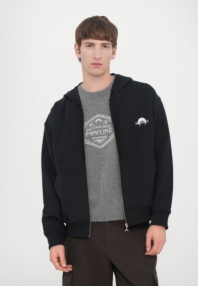 Felpa nera con zip e tasca frontale, indossata sopra un maglione grigio con un design grafico. Tessuto morbido, vestibilità casual, polsini a coste.