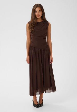 Vrouw met lang bruin haar draagt een mouwloze donkerbruine midi-jurk en zwarte hoge hakken, staand tegen een effen witte achtergrond.