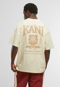 Crème-kleurige oversized t-shirt met een grote tan grafiek met "KANI" en decoratieve elementen op de achterkant, gemaakt van zacht katoen.