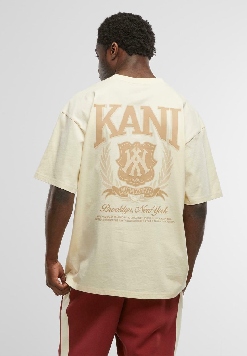 Crème-kleurige oversized t-shirt met een grote tan grafiek met "KANI" en decoratieve elementen op de achterkant, gemaakt van zacht katoen.