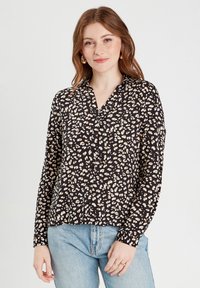 Chemise noire avec un motif léopard dans des tons beige et marron, manches longues, boutonnage à l'avant et col. Portée avec un jean bleu clair.