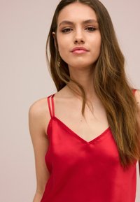 Rød satin camisole med tynde dobbelte stropper, der har en v-udskæring og glat tekstur. Model med langt hår poserer mod en neutral baggrund.
