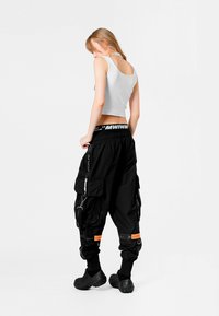 Pantalones cargo negros con múltiples bolsillos, detalles en naranja y cintura con cordón. Combinados con una camiseta de tirantes blanca corta y calzado negro.