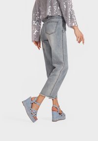 Jeans a gamba larga in denim con orlo sfrangiato abbinati a sandali con zeppa azzurri chiari, dotati di cinturini intrecciati e fibbie alla caviglia, su uno sfondo semplice.