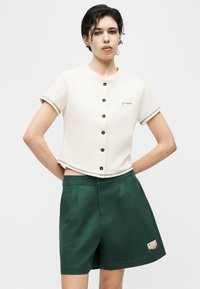 Νεαρό άτομο με κοντά σκούρα μαλλιά, που φοράει λευκό κοντομάνικο cropped πουκάμισο με κουμπιά και ψηλόμεσα σκούρα πράσινα σορτς, στέκεται με τα χέρια πίσω.