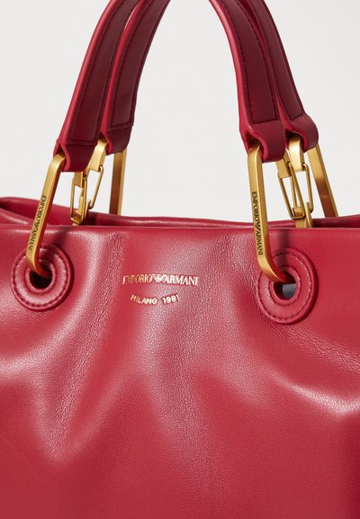Red leather handbag with gold hardware, embossed logo, and two short straps. Features smooth texture and circular grommet details.