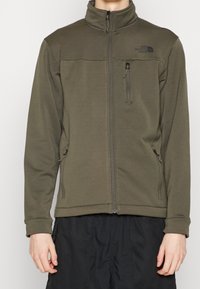 Fleecová bunda olivově zelené barvy s vysokým límcem, předním zipem a dvěma bočními kapsami. Na horní levé straně je černé logo. Měkká, texturovaná látka.