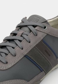 Zapatilla gris que combina materiales lisos y texturizados, con una distintiva raya azul y el logo de Geox en el lateral.