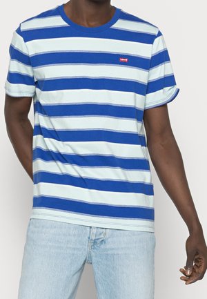 Mann trägt blaues, weißes und graues gestreiftes kurzärmeliges T-Shirt mit kleinem rotem Logo, kombiniert mit hellblauen Jeans.