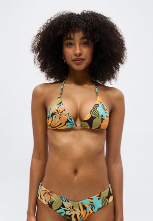 Mujer con cabello rizado que lleva un bikini colorido con estampado floral, de pie frente a un fondo gris claro y liso.