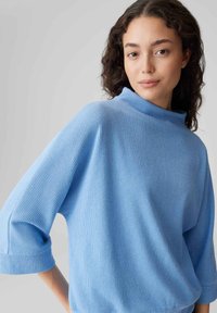 Hellblauer, gerippter Pullover mit Stehkragen und Dreiviertelärmeln. Hergestellt aus weichem Stoff, mit bequemem Schnitt und subtiler Textur.