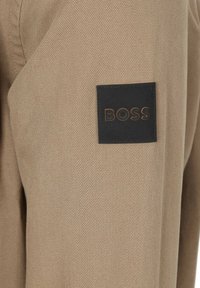 Beige stof met een gestructureerde weving; een zwart rechthoekig label met het woord "BOSS" in reliëf oranje letters aan de zijkant.