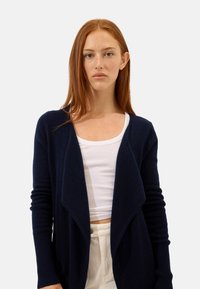 Mörkblå kofta i mjukt, strukturerat tyg med öppet fram, långa ärmar och ribbade ärmslut. Lagerdesignen ger visuell intress.