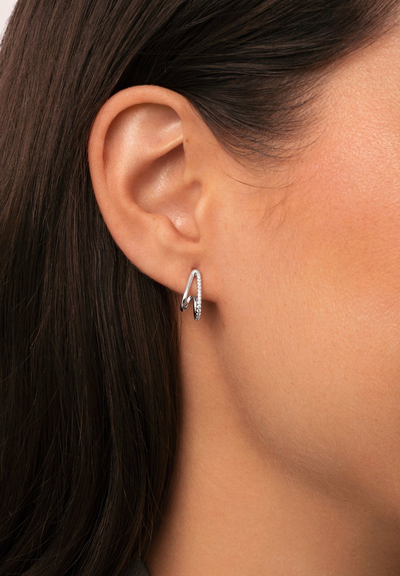 Orecchini d'argento con un design elegante e curvo, caratterizzati da una piccola sezione di pietre brillanti, indossati all'orecchio di un modello.
