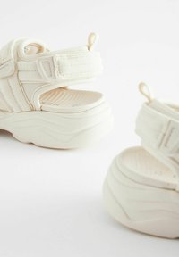 Sandales plates blanches avec fermeture à sangle, semelle intérieure texturée et forme arrondie. Fabriquées en matériau synthétique lisse.