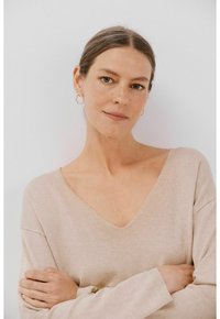 Beige V-hals sweater met lange mouwen, zachte breistekstuur en een relaxte pasvorm. Gouden creolen voegen een subtiele accent toe.