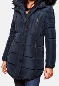 Marinblå vadderad jacka med huva, med svarta dragkedjefickor, vertikal sömnad och en lätt kurvad fåll för extra skydd.