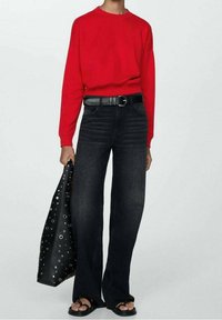 Person trägt ein rotes langärmeliges Oberteil, schwarze Jeans mit weitem Bein und schwarzem Gürtel, schwarze Sandalen und hält eine schwarze Handtasche mit Metallösen.