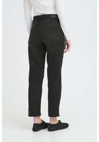 Osoba v čiernych jeansoch s rovnými nohavicami, bielom dlhom rukáve a čiernych slip-on topánkach stojaca na jednoduchom svetlom pozadí.