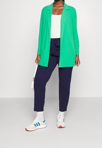 Zielona marynarka z długim rękawem, biały top na ramiączkach, granatowe spodnie zwężane z wiązaniem, białe trampki z niebieskimi akcentami, stojąca z małą torebką.