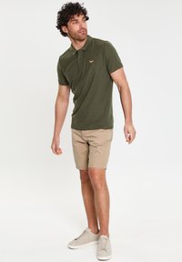 Polo verde in cotone con maniche corte, con un piccolo logo. Pantaloni corti beige, in tessuto leggero. Sneakers grigie chiare, design semplice.
