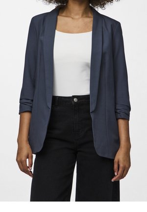 Blazer léger marine avec un col cranté, une ouverture frontale et des manches retroussées, associé à un top blanc et un pantalon noir taille haute.