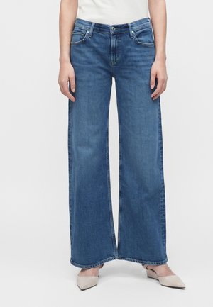 Personne portant un jean bleu à jambes larges et des chaussures beiges à bout pointu, debout sur un fond blanc uni.