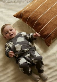 Grijze twee-delige outfit met spookpatronen, voorzien van een ronde halslijn en een elastische tailleband. Beige kussen met witte strepen op de achtergrond.