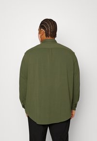 Oliwkowa koszula z długim rękawem, z zaokrąglonym dołem, z guzikami przy mankietach oraz detalem z tyłu w okolicy yoke, wykonana z lekkiego materiału.