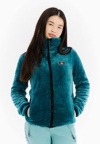 Teal fleece jack met hoge kraag, volledig ritssluiting en zwarte accenten. Voorzien van zijzakken en een patchlogo op de borst. Zachte textuur.