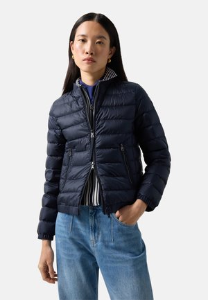 Kobieta ubrana w granatową pikowaną kurtkę na paskowanej koszuli i niebieskich dżinsach, z rękami swobodnie opuszczonymi wzdłuż ciała na jednolitym tle.