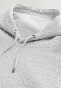 Sudadera blanca con capucha, con patrones texturizados y cordón ajustable. El forro interior es suave, con herrajes metálicos cerca del área del cuello.