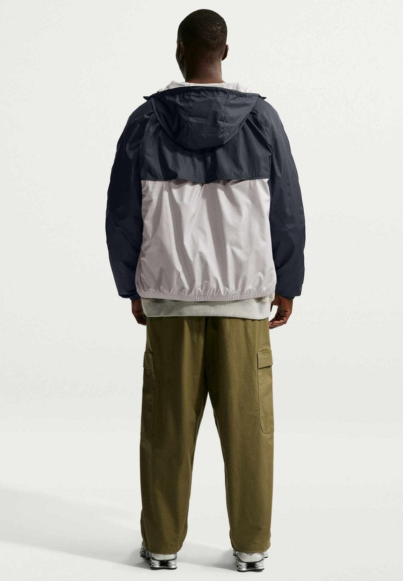 Man staat met zijn rug naar de camera gekeerd, draagt een tweekleurige navy en lichtgrijze capuchonjacket, olijfgroene cargobroek en witte sneakers op een effen achtergrond.