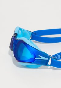 Blå svømmebriller laget av gjennomsiktig plast, med et glatt design, justerbart blått stroppe, og en buet linseform for klarhet.