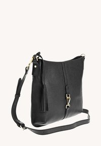 Sac en cuir noir à bandoulière réglable, avec un détail de pompon et un fermoir en métal doré sur un fond clair uni.