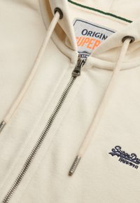 Cremfarbener Hoodie mit durchgehendem Reißverschluss, Kordelzügen und gesticktem Logo. Verfügt über einen weichen, strukturierten Stoff und einen kontrastierenden grünen Kragenfutter.