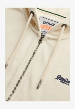 Cremefarvet hættejakke med fuld lynlås, snøre og broderet logo. Har et blødt, tekstureret stof og kontrasterende grøn kraveforing.