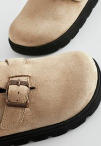 Chaussures en daim beige à bout rond, avec une sangle à boucle et une surface texturée, posées sur une semelle en caoutchouc noir.