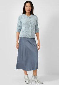 Lichtblauwe gebreide cardigan met ronde hals, zakken aan de voorkant en vijf glanzende knopen, gecombineerd met een grijze satijnen midi-rok en zilverkleurige enkellaarzen.
