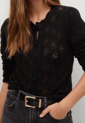 Femme portant un cardigan noir en maille boutonné avec un col festonné, rentré dans un jean noir taille haute avec une ceinture noire et une boucle dorée.