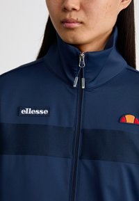 Mornarska zip-up jakna s visokim ovratnikom, vodoravnimi barvnimi blokami in izvezenim logotipom na prsih, izdelana iz gladkega materiala.